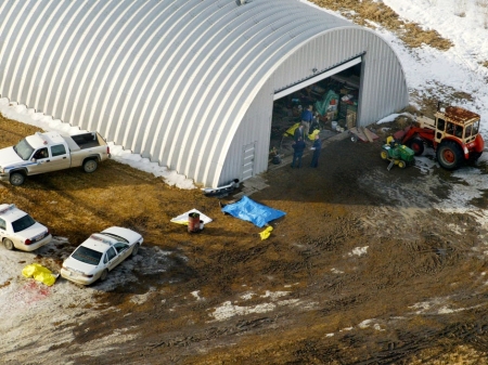 The tarped-over bodies of the four dead Mounties. One is to the rear of the police cruiser [note passenger side window shot out]; two appear to be under the blue tarp and the fourth under a yellow tarp just inside the quonset hut. Photo by Rick MacWilliam of the Edmonton Journal.